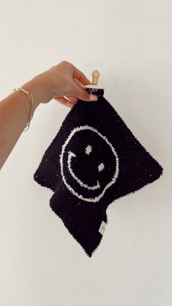 A hand holds up the "Lovey | Black & White Just Smile" by forever french baby, a black, textured washcloth featuring a cheerful white smiling face design. Made from microfiber feather yarn, the washcloth is displayed hanging from a small wooden peg against a plain white wall. The person wearing a silver bracelet appears to be using this versatile item as both a comforting lovey and practical accessory.
