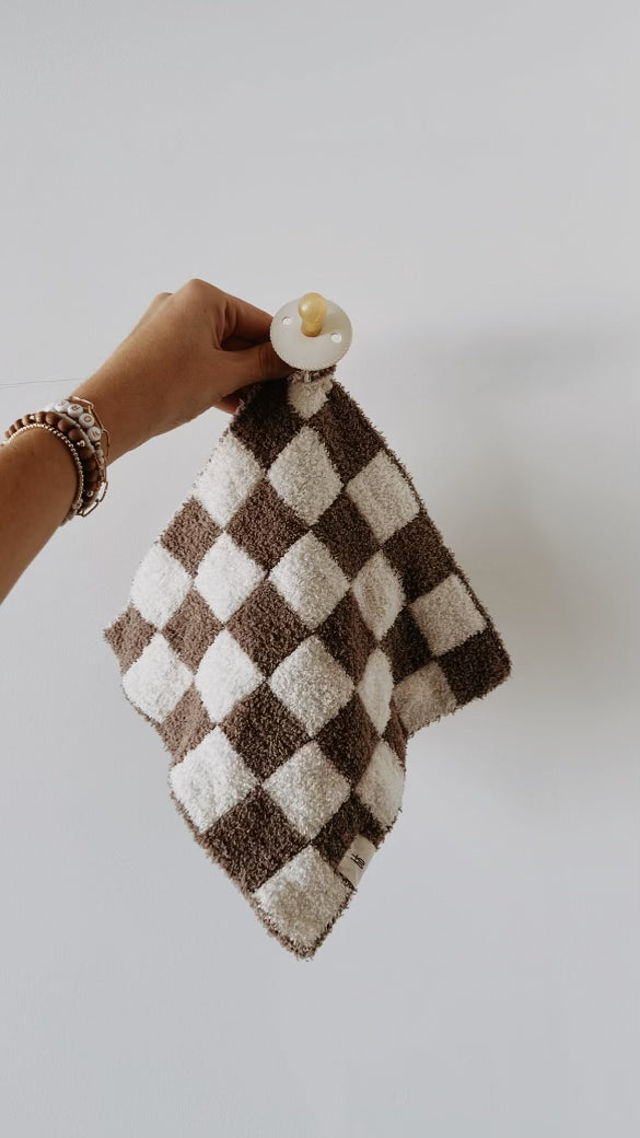 A hand holding up the Mushroom Checkerboard Lovey, a beige and brown classic checkerboard print cloth attached to a pacifier. The hand is adorned with a beaded bracelet, and the background is a plain white wall. This chic accessory by forever french baby adds timeless style to any baby's essentials.