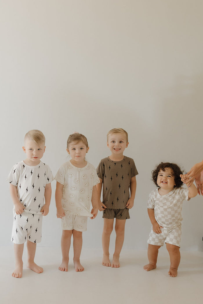 Four young children stand barefoot in a row against a light wall, each wearing playful, hypo-allergenic clothing. The child on the far right, smiling and holding an adult's hand, wears the "Bamboo Short Set | Brown & Black Lightning Bolt" by forever french baby.