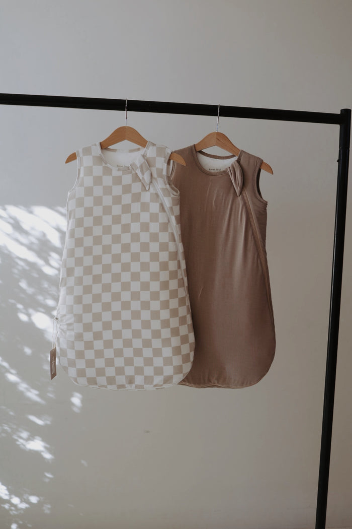 A Bamboo Sleep Sack by forever french baby hangs on a black clothing rack. One features a beige and white checkerboard design, while the grey version offers a solid look. Soft light and shadows highlight each wearable blanket against the plain wall.