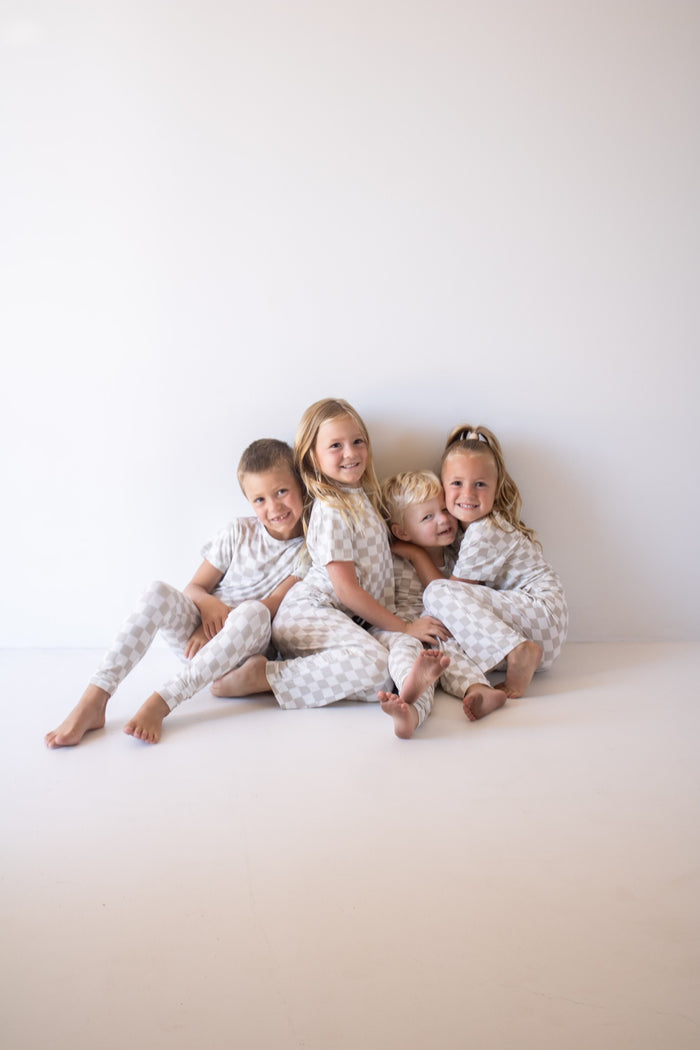 Four young children in matching forever french baby Child Boxy Tee Lounge Set | Checkerboard cuddle and smile together on a white floor against a plain white wall.