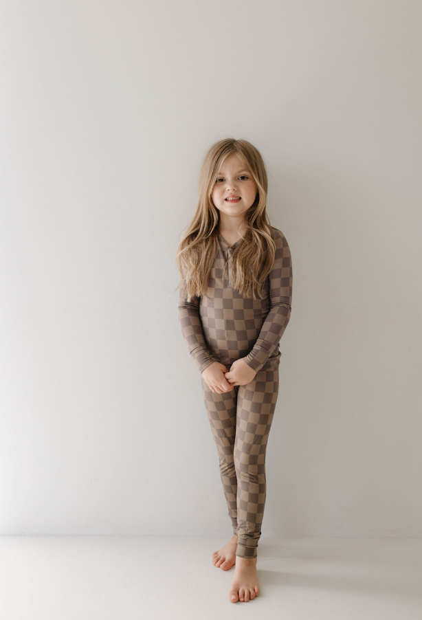 A young girl with long, blonde hair smiles as she stands against a plain white background. She is barefoot and wearing a matching set of breathable sleepwear in a faded brown checkerboard pattern by forever french baby, specifically the Bamboo Two Piece Pajama.