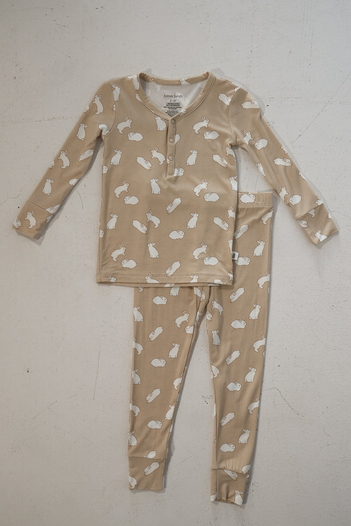The Bamboo Two Piece Pajamas | Bunnies by forever french baby features soft, hypoallergenic bamboo in beige with white bunny and cloud prints, a buttoned neckline, and matching pants, laid flat on a textured white surface.