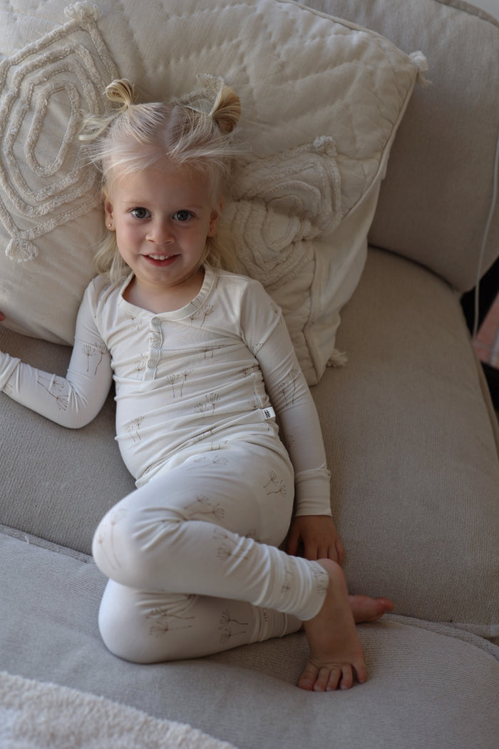 A young child with blonde hair in two buns smiles while lying on a light sofa, wearing forever french baby Bamboo Two Piece Pajamas in Dandelion print. A textured pillow rests behind her.