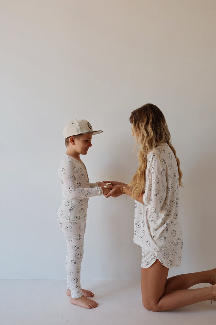 A woman kneels and holds hands with a smiling boy, both wearing forever french baby’s Bamboo Two Piece Pajamas | Green Smiles—matching white bamboo sleepwear with smiley faces, shown against a plain white wall.