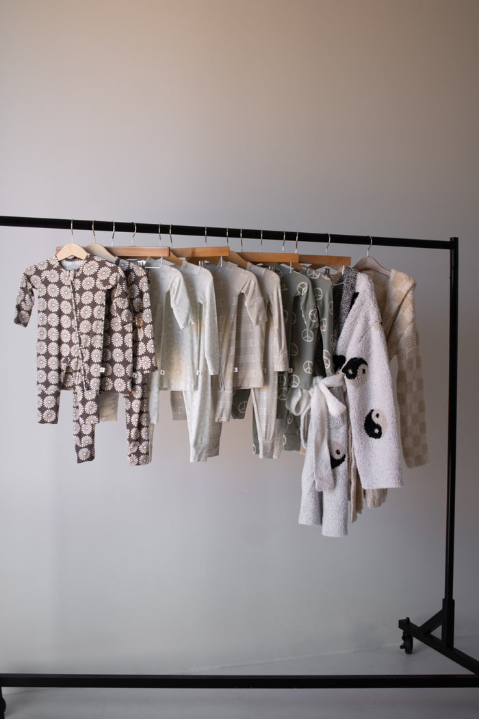 A clothing rack displays forever french baby’s Bamboo Two Piece Pajamas | Meow—children’s outfits in soft, neutral tones with playful prints—hanging neatly on wooden hangers against a plain background.