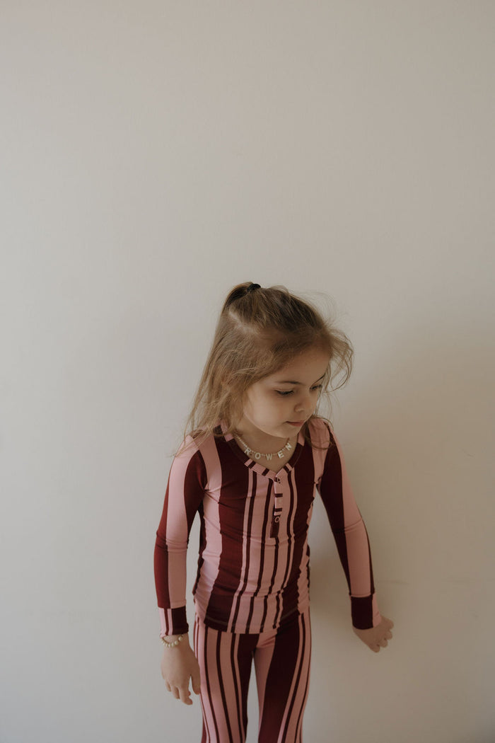 A young girl with light brown hair stands against a plain, light background wearing the forever french baby Bamboo Two Piece Pajamas in Sweetheart Stripes, featuring vertical maroon and pink stripes. She looks down and slightly to the side.