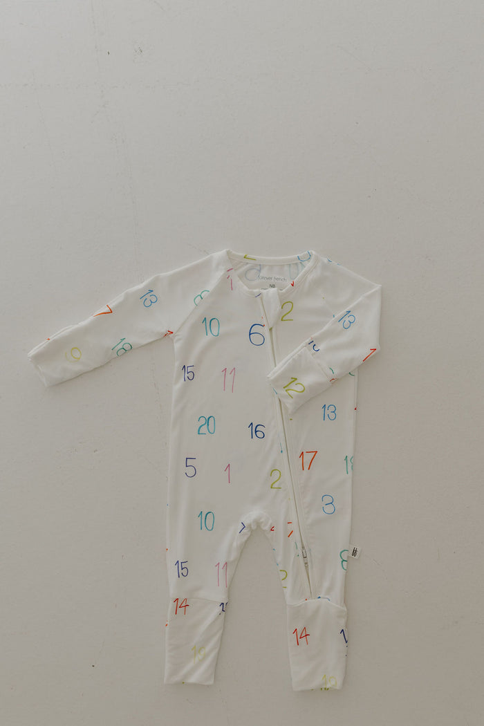 The Bamboo Zip Pajamas | Charli Counts by forever french baby is a white long-sleeve baby onesie featuring multicolored numbers. Crafted from hypo-allergenic bamboo fabric, it has a diagonal zipper from the neckline to the left foot, set against a light-colored background for a gentle design.