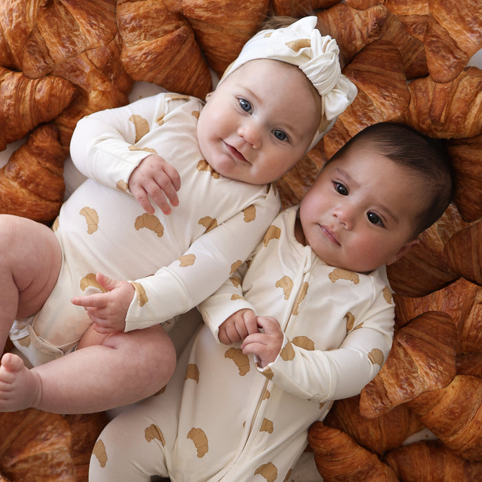 Two babies in matching forever french baby Bamboo Zip Pajamas | Croissants lie side by side on a croissant bed. One wears a bow headband, and both look content in their soft, hypoallergenic bamboo baby clothing.