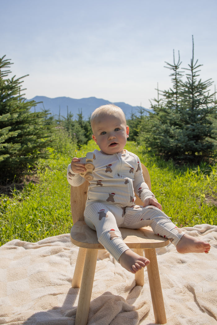 A baby dressed in the forever french baby Bamboo Zip Pajamas | Santa Teddy sits on a wooden chair outdoors, holding a wooden toy truck, surrounded by small pine trees and mountains in the background with sunlight streaming in.