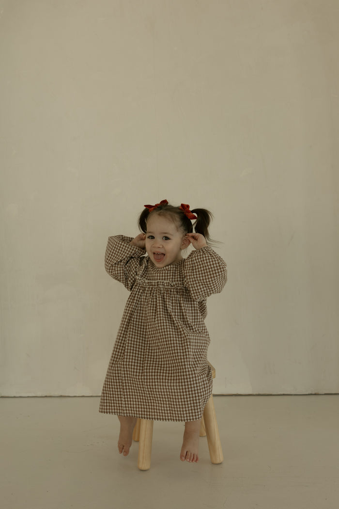 A young girl with pigtails and red bows sits barefoot on a stool, smiling playfully in the forever french baby Child Long Sleeve Dress | Honey Gingham against a plain beige background.