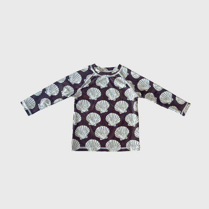 The Child Rash Guard | Seashells by forever french baby features long sleeves, a dark background, and a repeating white seashell pattern—ideal for kids' swimwear or summer outfits. Displayed flat on a light gray surface.