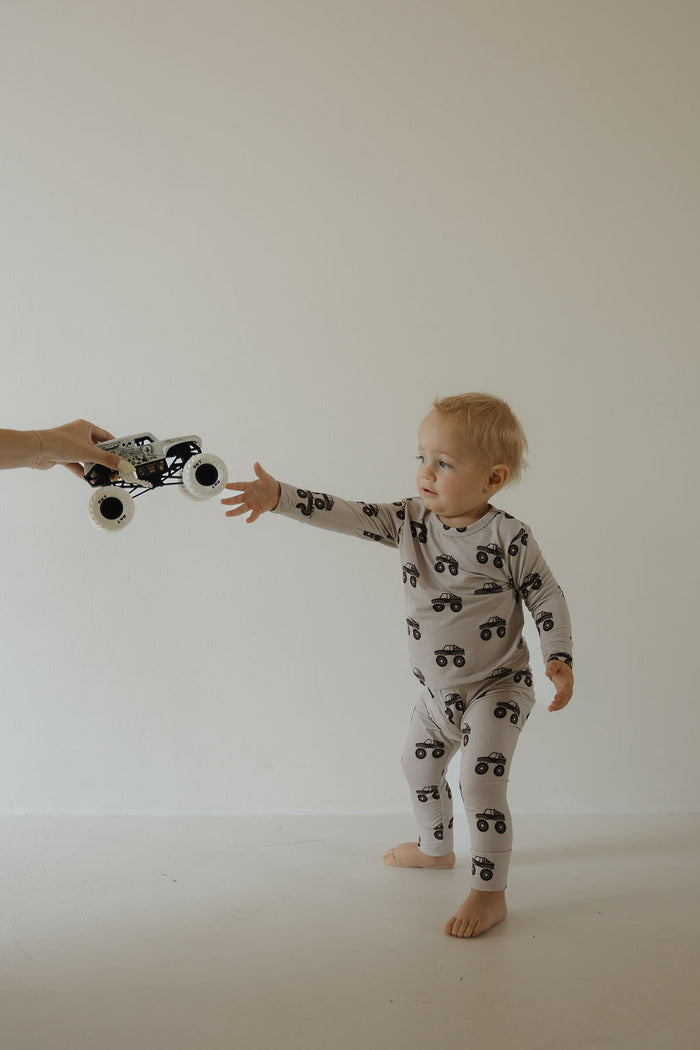 A toddler in the forever french baby Bamboo Footless Sleepy Time Set | Monster Truck stands barefoot, reaching for a toy monster truck from an adult against a plain, light background.