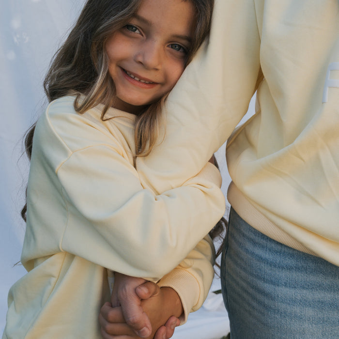 A young girl with long brown hair smiles while hugging an adult, both looking happy and close. She wears the forever french baby Child Sweatshirt | ff Yellow, paired with jeans, against a light background in matching family outfits.
