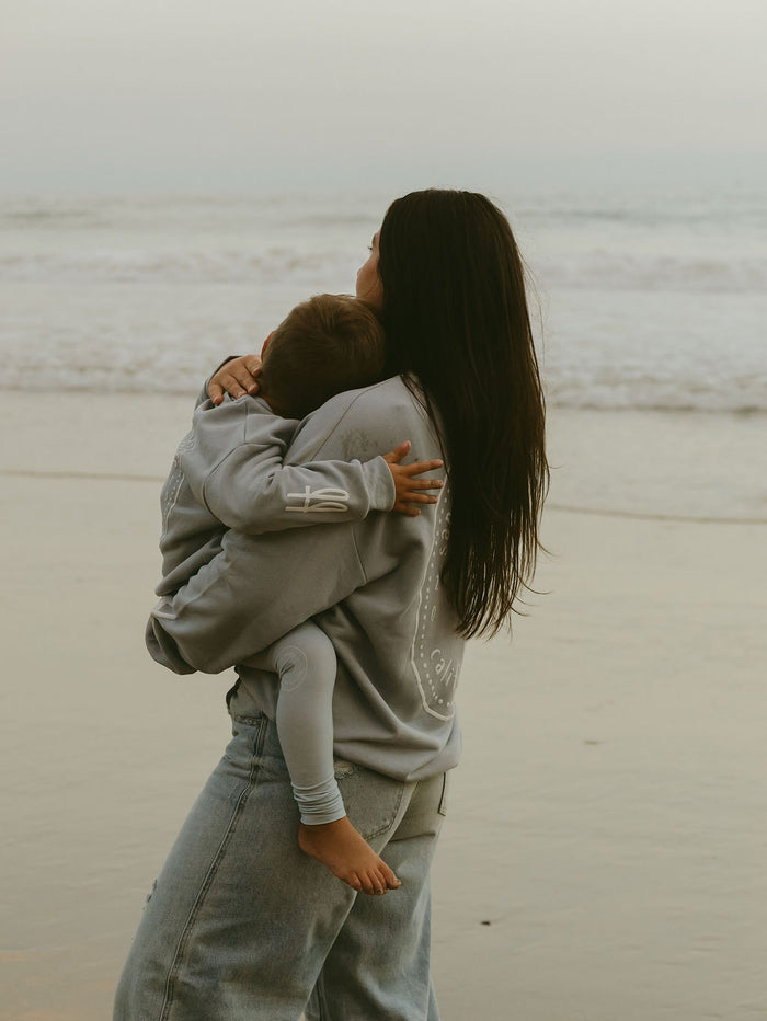A woman with long dark hair holds a child at the beach, both in jeans and the forever french baby Child Sweatshirt | San Clemente, as they gaze at the ocean—a perfect coastal keepsake moment.