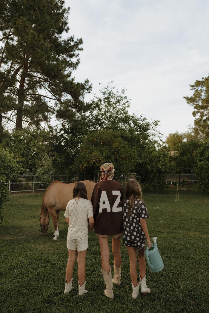 Three people stand on grass by a brown horse—one adult in an "AZ" shirt and two kids in boots, one holding a blue watering can—all wearing breathable forever french baby clothing like the Girl's Bamboo Short Set | With Love from AZ, amid trees outdoors.