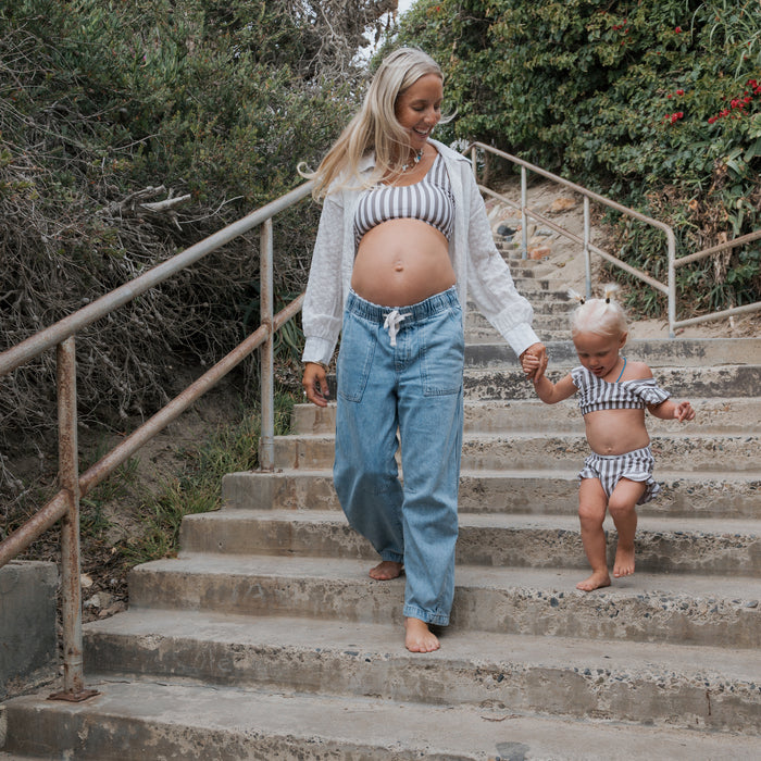 A barefoot woman and a young girl in matching forever french baby Olive Stripe Girl's Two Piece Swimsuits walk hand-in-hand down outdoor concrete steps surrounded by greenery.