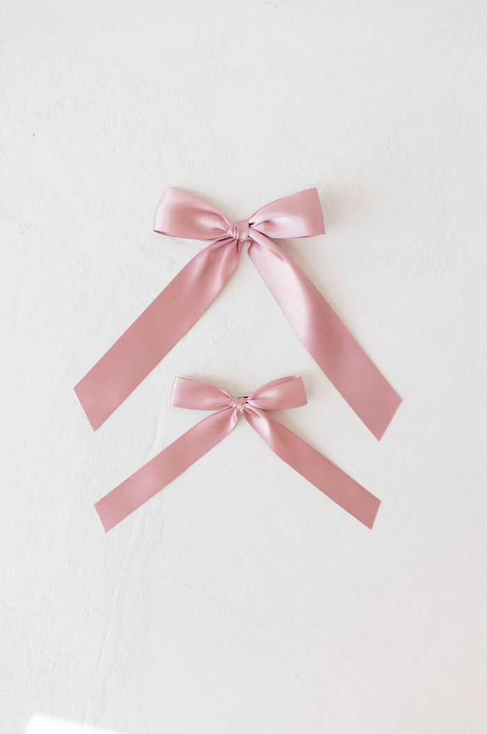 On a white surface, two satin hair clips from forever french baby are elegantly laid out. The top Hair Bow Clip | Satin showcases a larger bow with longer trailing ribbons, while the bottom one features a smaller bow with shorter ribbons. Both pink bows are neatly tied and exhibit a shiny, smooth appearance.
