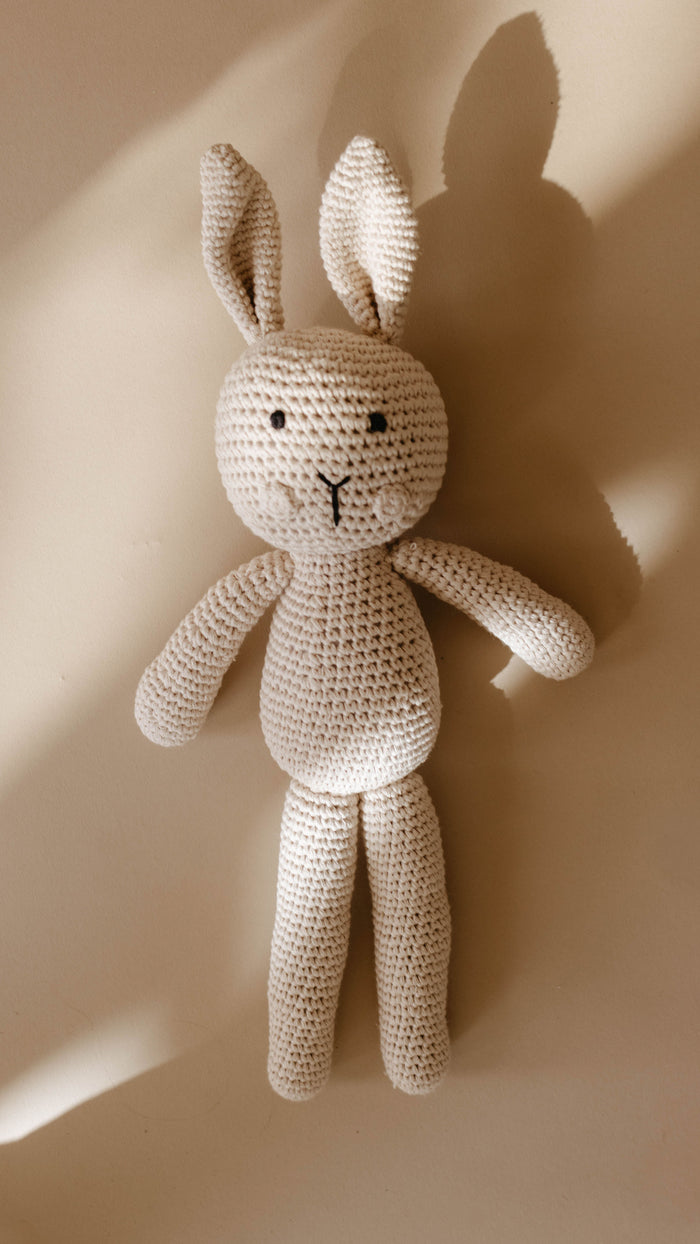 Honey the Bunny, a hand-crocheted knit doll from forever french baby, lies on a light beige surface. Crafted with light beige yarn, this charming stuffed bunny features long ears, small black eyes, and a stitched mouth. It casts a soft shadow and exudes the timeless charm of a Forever French Baby treasure.