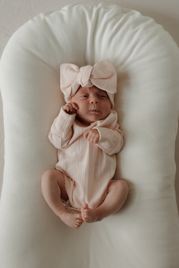 A baby wearing the forever french baby Long Sleeve Snapsuit | Ballerina in pale pink pointelle cotton, paired with a large matching bow headband, lies on a white cushioned surface, looking up with one hand near their face and legs slightly bent.