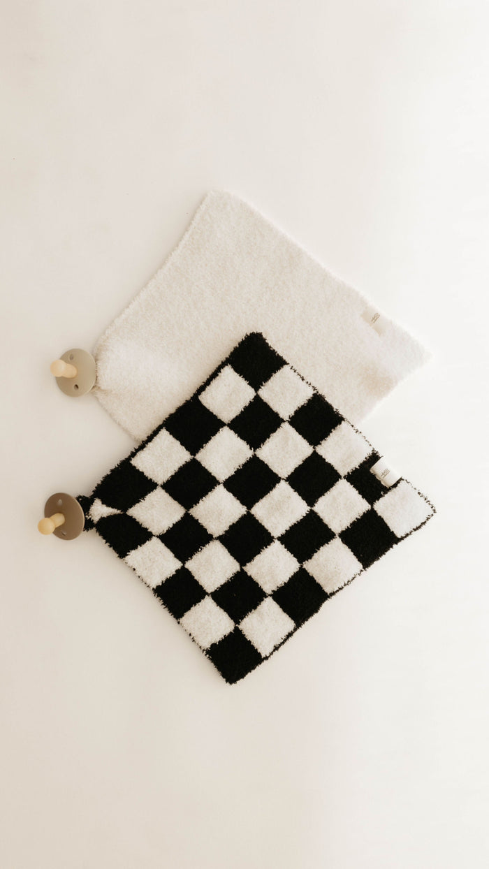 Two textured rectangular mats with small wooden bear-shaped knobs are displayed against a white background. One mat, crafted from microfiber feather yarn, is plain white, while the other is the "Lovey" by forever french baby, featuring a striking black-and-white checkerboard pattern.