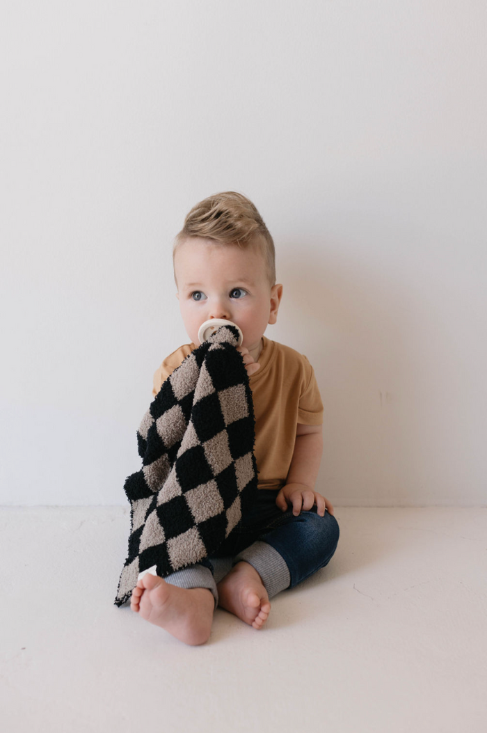 A small child with short blond hair sits against a white wall, wearing a tan shirt and blue jeans. The child is holding the Lovey | Checkmate Checkerboard blanket from forever french baby and has a pacifier in their mouth, looking slightly to the side.