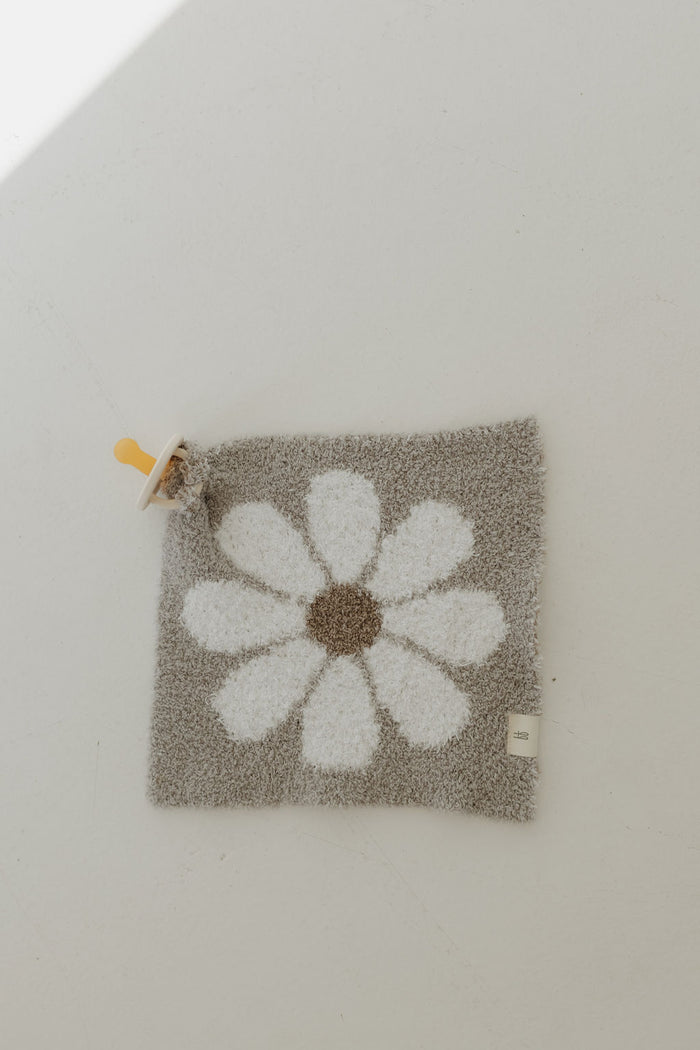 On a light surface lies the "Lovey | Darlin'" by forever french baby, a soft square gray baby blanket crafted from microfiber feather yarn, featuring a white daisy pattern with brown centers and a yellow pacifier attached to one corner.