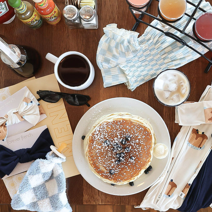 A plate of pancakes topped with blueberries and powdered sugar sits on a table for the Lovey | Morning Check by forever french baby, surrounded by coffee, juice, utensils, napkins, sunglasses, and a bow tie.