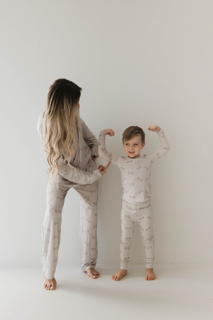 An adult and a child wearing matching Adult Long Sleeve Bamboo Pajamas | Arm Day by forever french baby stand against a plain white background. The child faces the camera, smiling, and flexing their arms proudly while the adult, standing next to them, looks towards the child. Both sets of pajamas feature a subtle dumbbell weights graphic pattern.