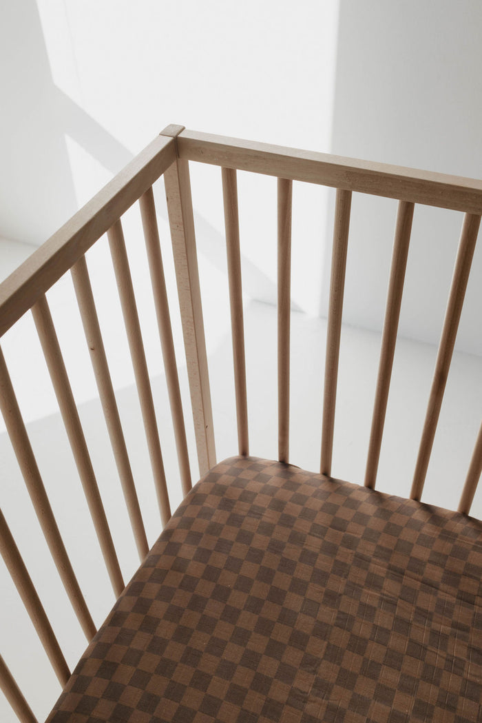 A wooden chair with vertical slats and a brown checkered cushion, similar to the Muslin Crib Sheet | Faded Brown Checkerboard by forever french baby, is bathed in natural light. The sunlight casts soft shadows on the chair, revealing its simple yet elegant design.