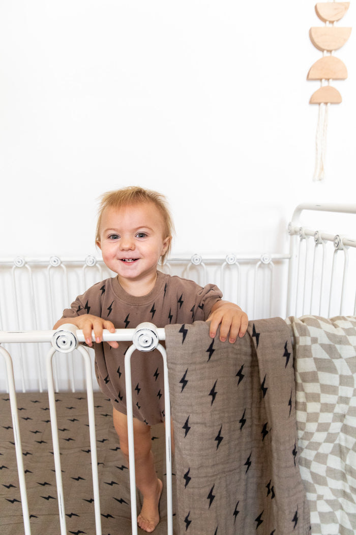 A smiling toddler with light hair stands inside a white crib, holding onto the rail. The crib is adorned with a Muslin Crib Sheet in a lightning bolt pattern by Forever French Baby and complemented by a checkered muslin cotton blanket. In the background, there's a white wall decorated with a hanging wooden mobile.