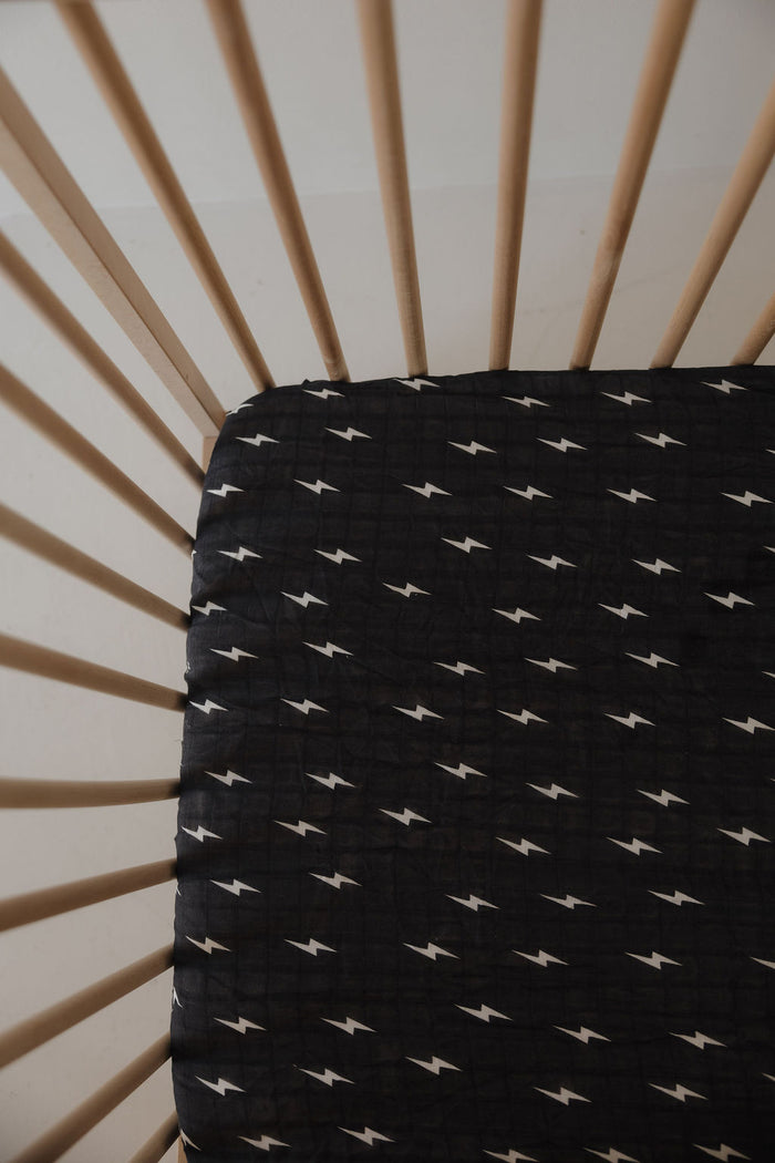 A wooden crib with vertical slats and a forever french baby Muslin Crib Sheet | Midnight Bolt—breathable black muslin with a white lightning bolt pattern—for stylish baby bedding.