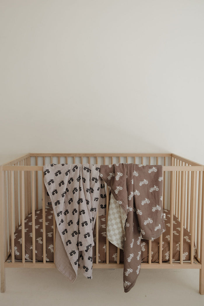 A light wooden crib with a brown fitted sheet features a forever french baby Muslin Quilt | Monster Truck, adding charm to the nursery against a white wall.