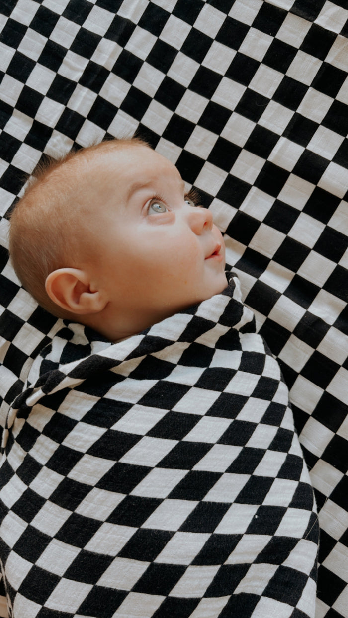 A baby wrapped in a forever french baby Muslin Swaddle | Black & White Checkerboard lies on a matching checkered surface, gazing upward with wide eyes.
