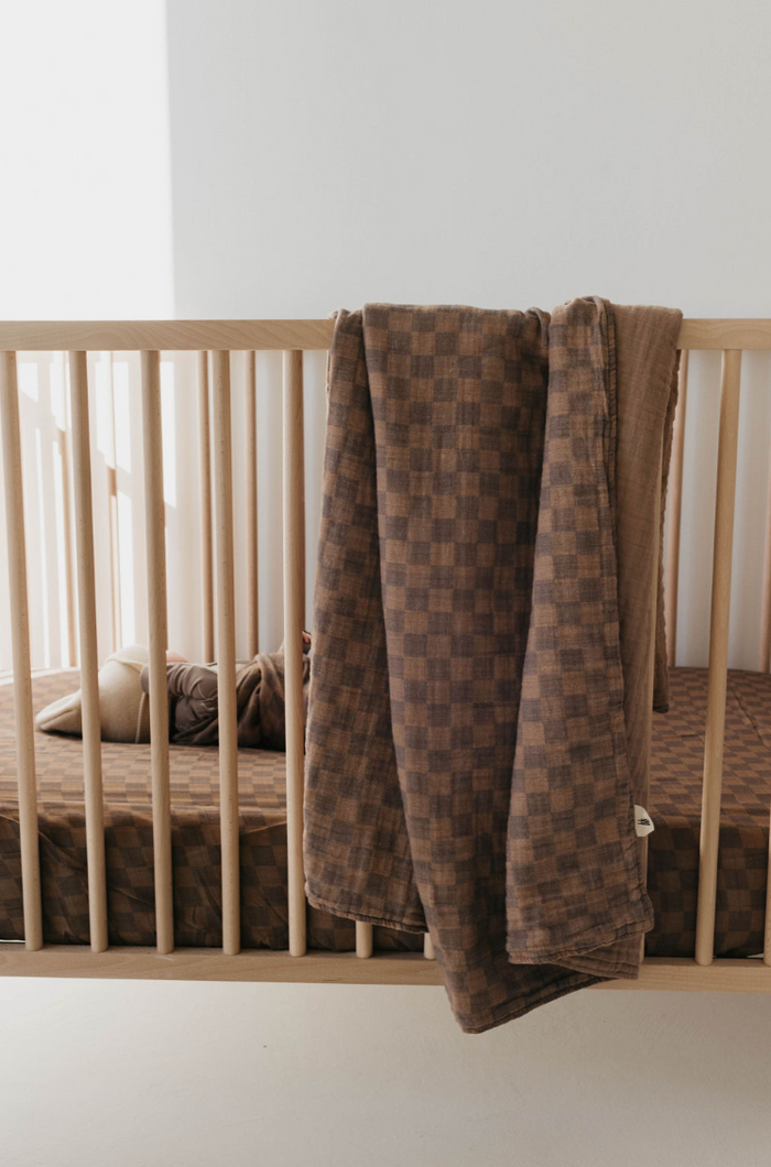 A wooden baby crib with a forever french baby Muslin Swaddle in Faded Brown Checkerboard draped over the side. Inside the crib, matching checkered bedding complements the swaddle, while a few soft toys rest on the mattress. Sunlight streams softly into the room, creating a play of light and shadows.