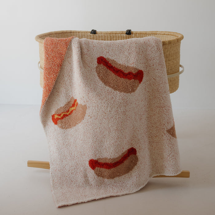 The forever french baby Plush Blanket | Hot Dogs—a beige knit microfiber, Oeko-Tex certified—features playful hot dog designs and is shown draped over a woven basket against a white background.