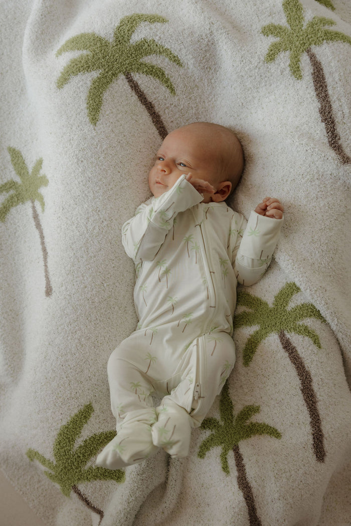 A baby in a white onesie with a palm tree print lies on the forever french baby Plush Blanket | Palm, a soft, Oeko-Tex certified microfiber blanket decorated with large green and brown palm trees.