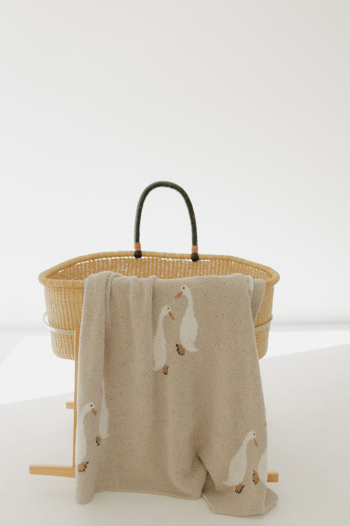 A woven basket with dark handles sits on a wooden stand. The forever french baby Plush Blanket | Silly Goose, Oeko-tex certified and decorated with white ducks, drapes over the edge of the stand in a bright minimalist setting.