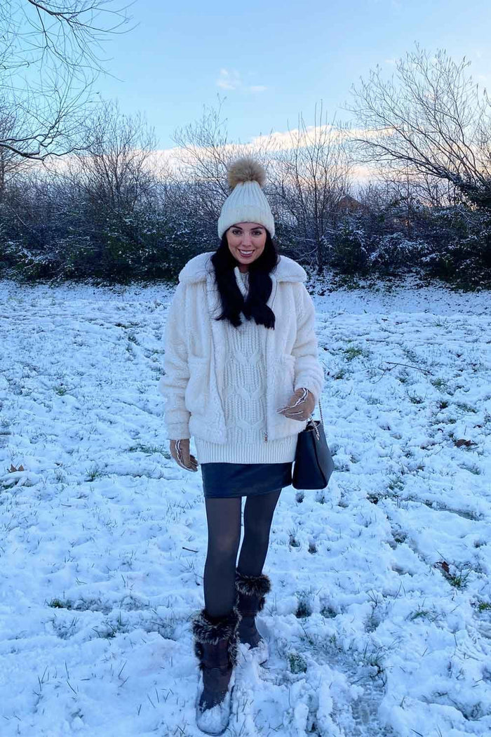 Model wearing a white knit sweater with sheer fleece lined tights and hat standing in a snowy field.