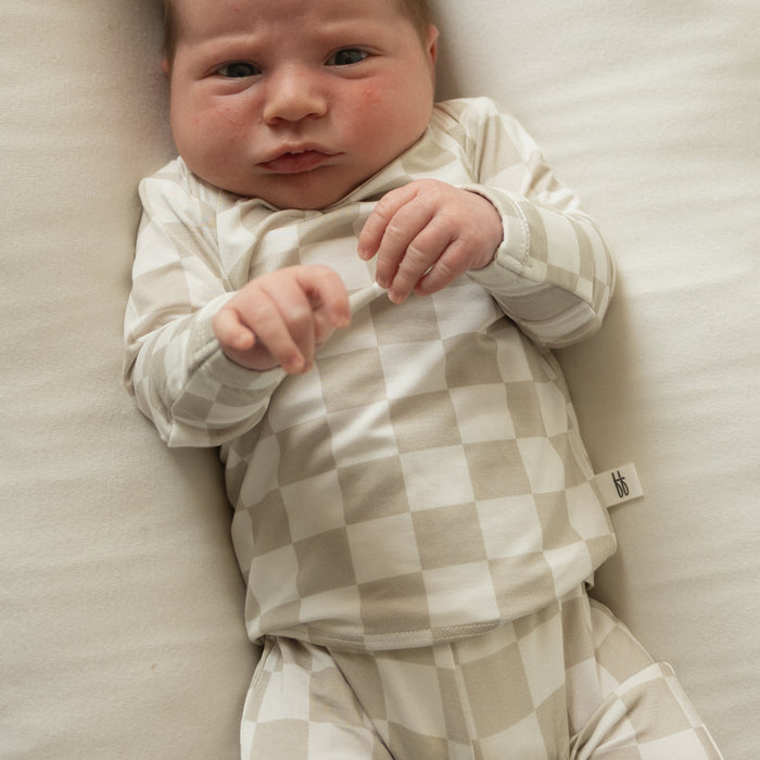 A newborn rests on a cream blanket, wearing the forever french baby Sleepy Time Set | Checkerboard in beige and white, gazing up calmly with gently clenched fists.