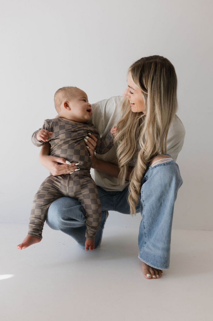 A woman with long blonde hair, dressed in a white t-shirt made of breathable bamboo fabric and blue jeans, kneels and laughs with a baby. The baby, wearing the Sleepy Time Set in Faded Brown Checkerboard by Forever French, looks up at the woman with a smile. The background is plain white.