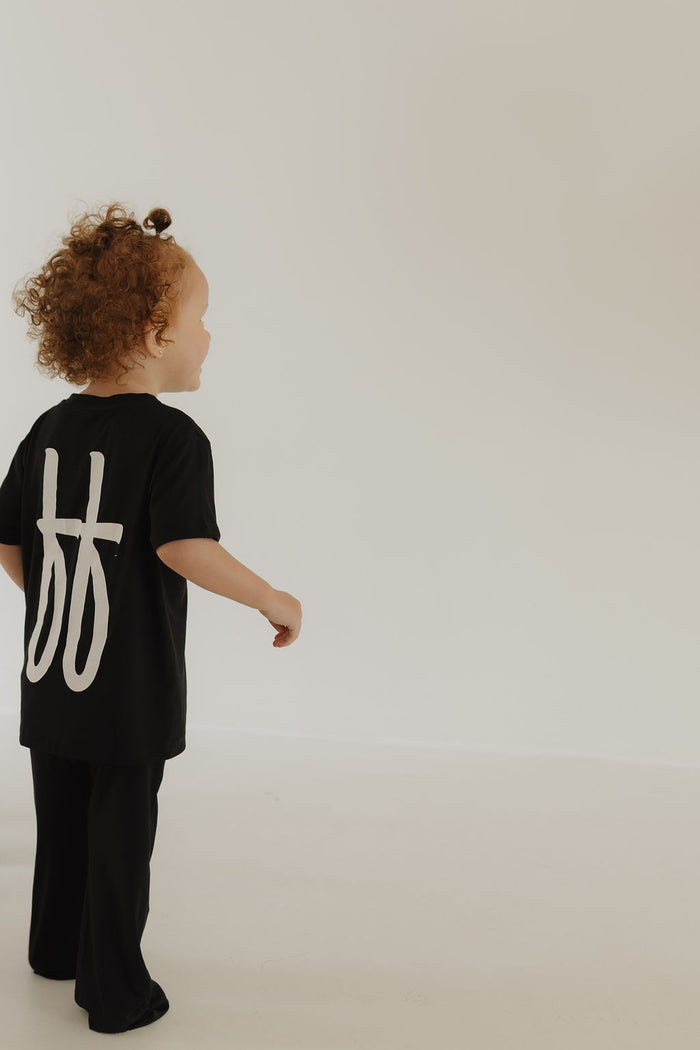 A young child with curly hair stands against a plain, light-colored background, looking to the side while wearing the "Toddler Short Sleeve Bamboo Lounge Set | Black ff Signature" from forever french baby, featuring a large white design on the back of the shirt.