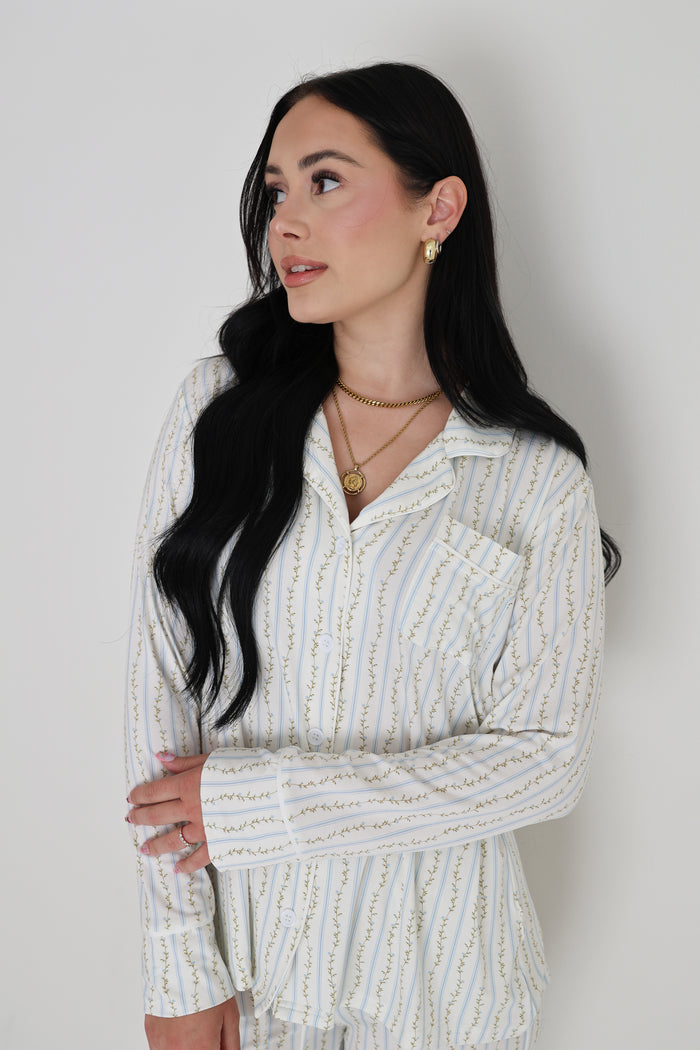 Woman with long black hair wearing the forever french baby Women's Bamboo Button Up Lounge Set in Dainty Vines, featuring striped pajamas with a pocket, layered gold necklaces, and gold earrings, standing against a plain white background.