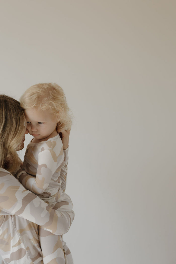 A woman holds a young blonde child close, both wearing matching forever french baby Women's Bamboo Pajama | Camo 2.0 sets in a beige and cream pattern, against a plain light background.