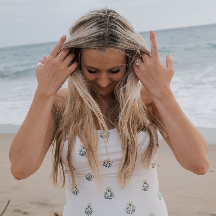 A woman with long blonde hair stands on a sandy beach, adjusting her hair in the Forever French Women's One Piece Swimsuit | Marigold, featuring a white base with green and blue florals, as the ocean provides a serene backdrop.