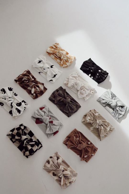 A set of 14 adjustable Bamboo Head Wraps from forever french baby, featuring the Faded Brown Checker design among others, are neatly arranged on a white surface. These headbands, made from soft bamboo material, showcase various patterns including polka dots, geometric shapes, stripes, and animal prints in shades of browns, blacks, whites, and grays.