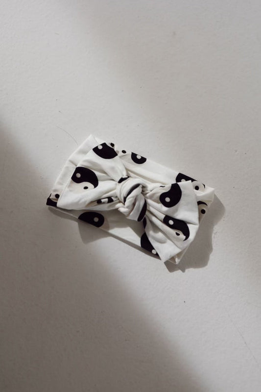 A Forever French Baby Bamboo Head Wrap in black and white yin-yang pattern, featuring a knotted bow in the center, is placed on a smooth, light-colored surface. This elegant piece seamlessly integrates charm and style.