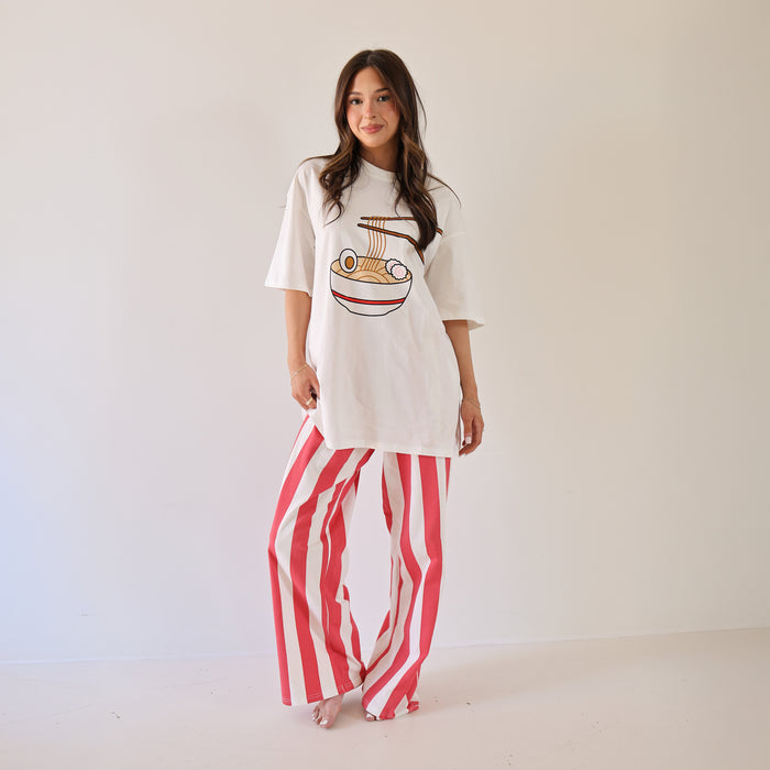A woman smiles barefoot against a white wall, wearing a white tee with a burger graphic and forever french baby Women's Wide Leg Pants | Tokyo Stripe—red-and-white vertical stripes and an elastic waistband. Her long brown hair flows freely.