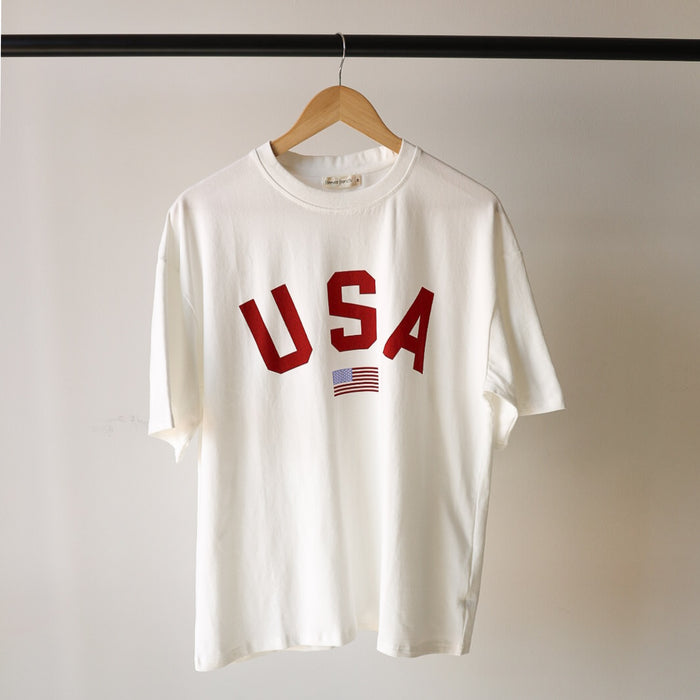 The forever french baby Adult Cotton Tee | USA is a 100% cotton unisex t-shirt on a wooden hanger, featuring bold red "USA" lettering with a small American flag graphic below.