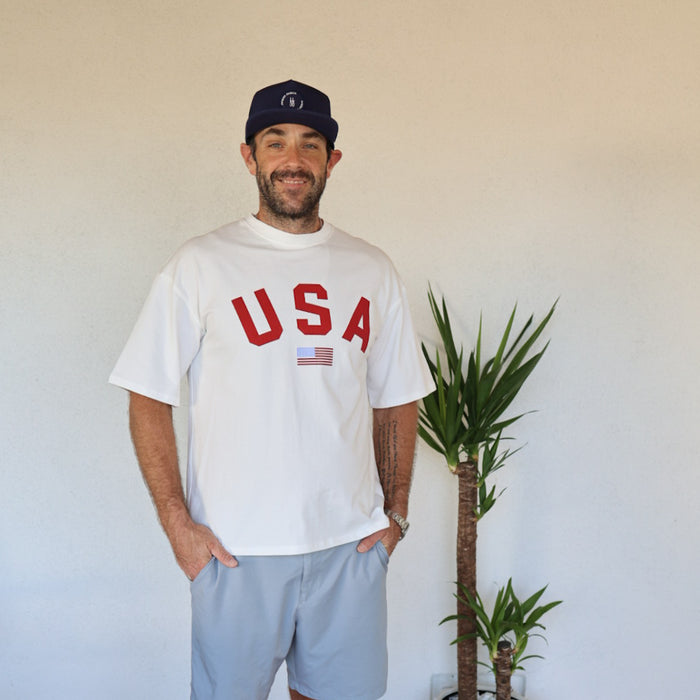 A smiling man in a blue hat and gray shorts stands indoors by a green plant, wearing the forever french baby Adult Cotton Tee | USA—an all-cotton unisex shirt with "USA" and an American flag—against a plain light wall.